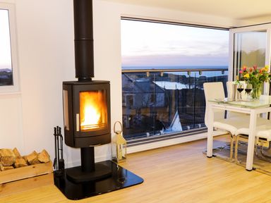 A room with a modern wood-burning stove next to a glass dining table with chairs and a sea view at Sunset View in Porth