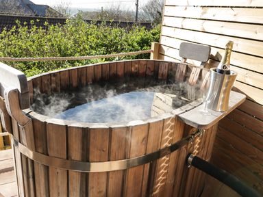 An outdoor wooden hot tub with steam rising next to a metal bucket holding a bottle of champagne and a glass on a wooden deck at Sunset View in Porth