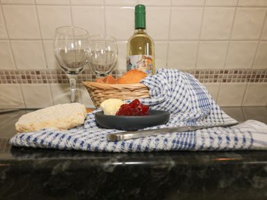 A kitchen countertop with a blue and white cloth holding bread, butter, and jam with a basket of bread and a bottle of wine with two wine glasses in the background at Lodge 22 Bodmin Holiday Park Cornwall Lanivet
