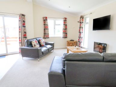 A living room with two black sofas a wooden coffee table a wall-mounted TV and windows with patterned curtains at Callow Lodge 22 in Beaconsfield Holiday Park