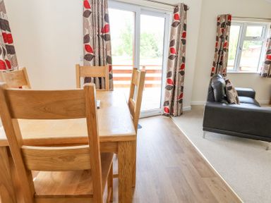 A dining area with wooden chairs and table next to a sliding glass door with leaf patterned curtains beside a living room with a black sofa and matching curtains at Callow Lodge 22 in Beaconsfield Holiday Park