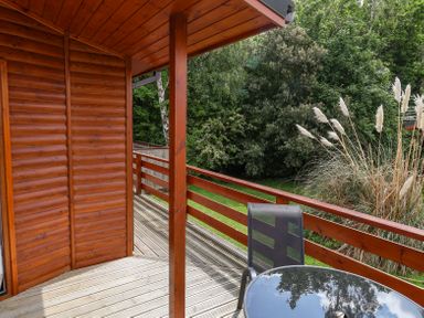 A wooden balcony with a glass table and chair overlooking green trees and plants at Callow Lodge 22 Beaconsfield Holiday Park