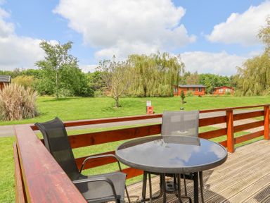 An outdoor deck with two black chairs and a round glass table overlooking grass trees and wooden cabins at Callow Lodge 22 Beaconsfield Holiday Park