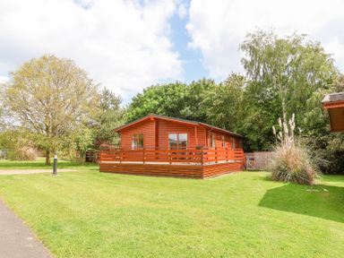 A wooden lodge with a fenced porch on a grassy area with trees at Callow Lodge 22 Beaconsfield Holiday Park
