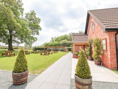 An outdoor area with picnic tables on grass and potted plants along a walkway next to a brick building at Callow Lodge 22 in Beaconsfield Holiday Park