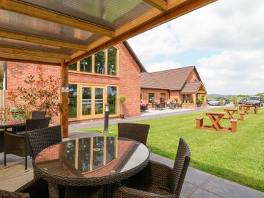 An outdoor patio area with wicker chairs and tables next to a brick building with large windows and wooden picnic tables on the grass at Callow Lodge 22 Beaconsfield Holiday Park