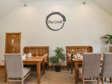 A dining area with wooden tables and chairs with beige upholstery and wall art that says The Croft at Callow Lodge 22 in Beaconsfield Holiday Park