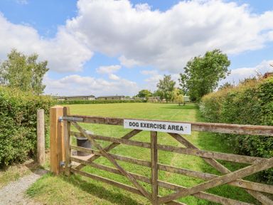 A grassy dog exercise area enclosed by a wooden gate with a sign and surrounded by hedges and trees at Callow Lodge 22 in Beaconsfield Holiday Park