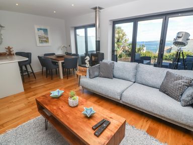 A living room with a gray sofa and wooden coffee table near dining table and large windows overlooking the sea at Seafield in Carbis Bay near St Ives