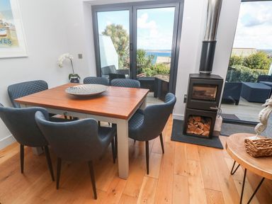 A dining area with a wooden table surrounded by six blue chairs near a stove and glass doors opening to an outdoor seating area with a view at Seafield in Carbis Bay near St Ives