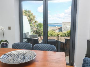 A dining area with a wooden table and blue chairs next to glass doors opening to a patio with wicker furniture and sea view at Seafield in Carbis Bay near St Ives