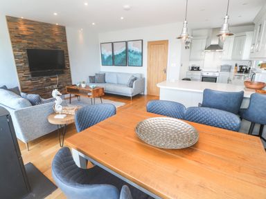 An open plan living room and kitchen with blue chairs a wooden dining table light grey sofas and a wall mounted television at Seafield in Carbis Bay near St Ives