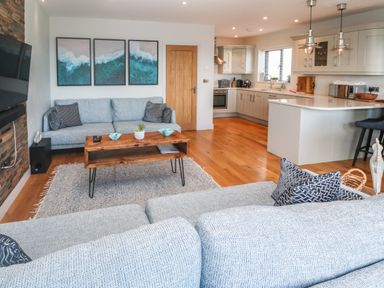An open living room and kitchen with a blue sofa wooden coffee table paintings on the wall and kitchen cabinets at Seafield in Carbis Bay near St Ives