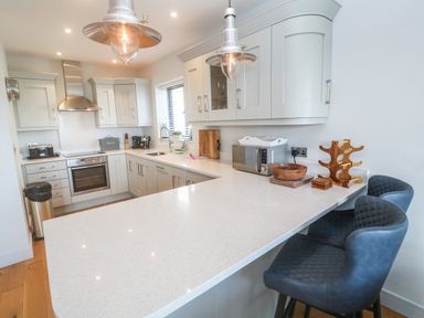 A kitchen with white cabinets a stainless steel oven and hood a white countertop and two blue bar stools at Seafield in Carbis Bay near St Ives