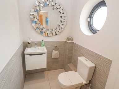 A small bathroom with a toilet, a wall-mounted sink, a round decorative mirror, and a porthole window at Seafield in Carbis Bay near St Ives