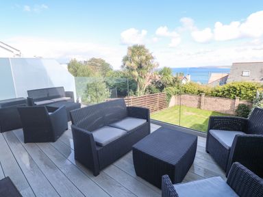 An outdoor patio with black wicker furniture and a glass railing overlooking a grassy yard and ocean view at Seafield in Carbis Bay near St Ives