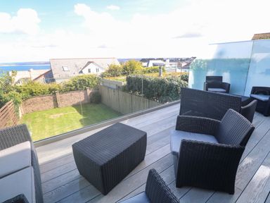 An outdoor balcony with black wicker furniture overlooking a garden and houses with a view of the sea at Seafield in Carbis Bay near St Ives