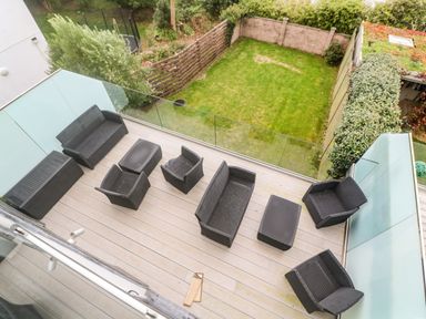 An outdoor patio with dark wicker furniture including chairs sofas and tables enclosed by glass panels overlooking a fenced garden