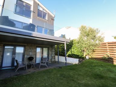 A backyard with grass a patio area with chairs a small table and a barbecue in front of a two story house with glass balcony at Seafield in Carbis Bay near St Ives