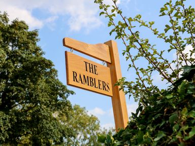 A wooden sign reading The Ramblers hanging outdoors near green foliage and trees at The Ramblers' Annex in Mattishall