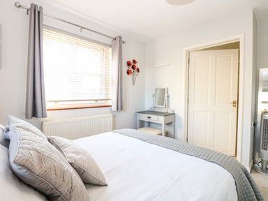A bedroom with a bed, pillows, a window with blinds and curtains, a small dressing table with a mirror, and a door at The Ramblers Annex in Mattishall