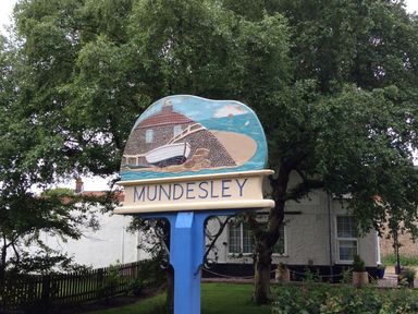A wooden sign with a depiction of a boat and beach with the word Mundesley in a garden with trees and a house in the background at 20 Siesta Mar in Mundesley