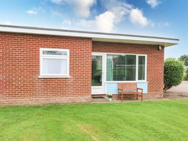 A brick building with a white door and windows a wooden bench and a potted plant on grass at 20 Siesta Mar in Mundesley