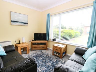 A living room with two black sofas a blue rug wooden tables a television and a large window with blue curtains at 20 Siesta Mar in Mundesley