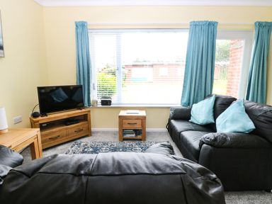 A living room with black leather sofas and blue cushions a wooden TV stand with a television a small wooden side table and blue curtains at 20 Siesta Mar in Mundesley