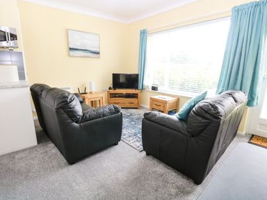 A living room with two black leather sofas a wooden TV stand and side tables and a large window with blue curtains at 20 Siesta Mar in Mundesley
