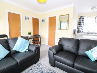 A living room with black sofas and blue cushions a table with chairs and a kitchen in the background at 20 Siesta Mar in Mundesley