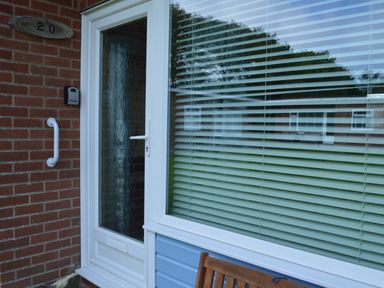 A white door with a glass panel next to a large window with closed blinds on a brick wall with a wooden chair in front at 20 Siesta Mar in Mundesley