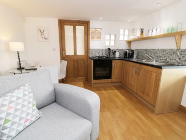 A small kitchen area with wooden cabinets a stove a sink and a shelf with cups and containers a wooden door and a gray sofa with a patterned cushion at The Annex @ Garden House Prudhoe near Newcastle