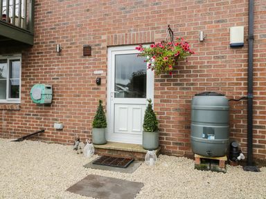 A brick house exterior with a white door flanked by two potted plants a hanging basket with pink flowers and a rain barrel at The Annex @ Garden House in Prudhoe near Newcastle