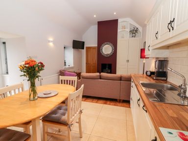 A kitchen and dining area with a wooden table and chairs a vase with flowers on the table a sofa TV and cupboards at Laharn Cottage in Killorglin County Kerry