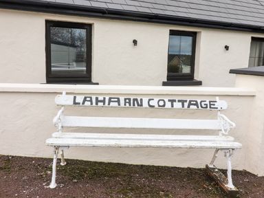 A white bench with LAHARN COTTAGE written on it outside a cream building with two windows at Laharn Cottage in Killorglin County Kerry