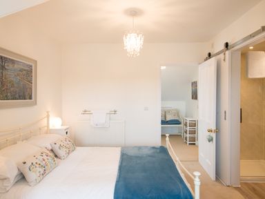 A bedroom with a bed with white covers and blue blanket next to a nightstand and an open door leading to another room and a bathroom at Spiral View in Perranporth