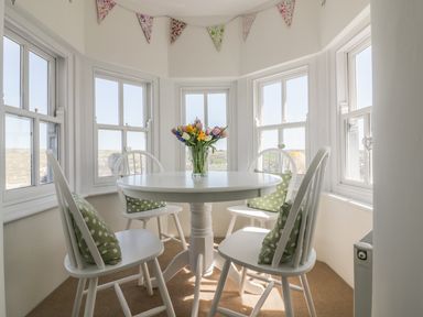 A round white dining table with four white chairs and green polka dot cushions in a room with multiple windows and a floral garland at Spiral View in Perranporth