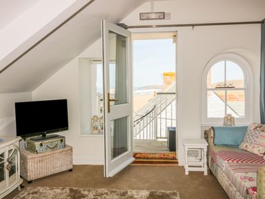 A room with a patchwork sofa and TV near a door opening to a balcony overlooking a beach at Spiral View in Perranporth