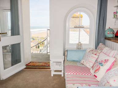 A seating area with a patterned sofa and cushions next to a window and an open door leading to a balcony overlooking the beach at Spiral View in Perranporth