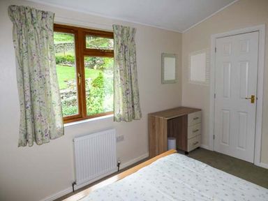 A bedroom with a wooden window and floral curtains a radiator a wooden desk with drawers a white door and part of a bed at South Lodge in Kendal