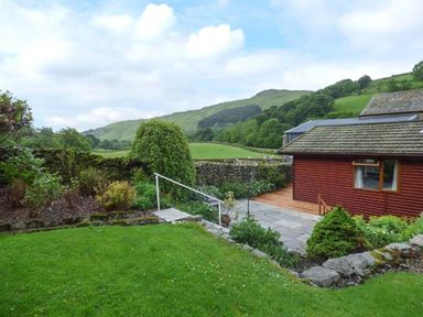 A garden with a green lawn shrubbery and stone wall next to a red building with hills in the background at South Lodge in Kendal