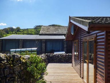 A wooden cabin with a deck and stone wall surrounded by greenery and hills at South Lodge in Kendal