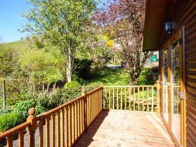 A wooden deck with a railing attached to a house overlooking a garden of trees and bushes at North Lodge in Kendal