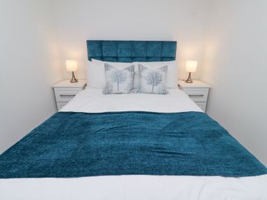 A bedroom with a bed with blue headboard two white nightstands with lamps and pillows at 6 Mill Wharf in Berwick-upon-Tweed