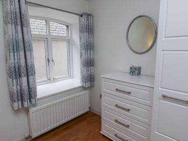 A small room with a window with patterned curtains a white radiator below the window a white chest of drawers with a round mirror above it and a tissue box on top at 6 Mill Wharf in Berwick-upon-Tweed