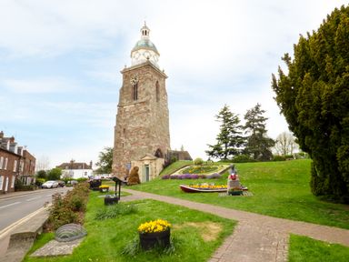 A stone clock tower with a green dome on a grassy hill next to a path and flower beds at Garden View in Malvern