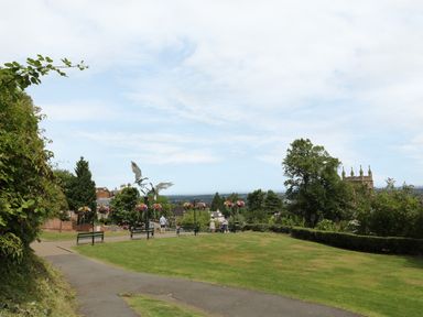 A park with benches flower pots and a metal sculpture of birds with buildings and trees in the background at Garden View in Malvern