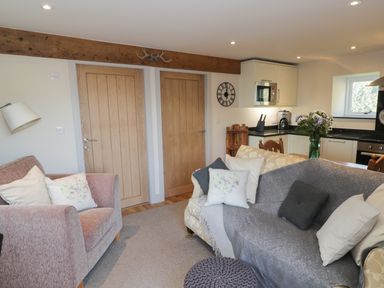 A living area with a pink upholstered armchair, two wooden doors, a gray sofa with cushions, a kitchen with white cabinets and appliances, and a wooden dining table with flowers at The Granary Loft in Bardsea