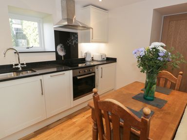 A kitchen with white cabinets black countertops a stove and a wooden dining table with flowers at The Granary Loft in Bardsea
